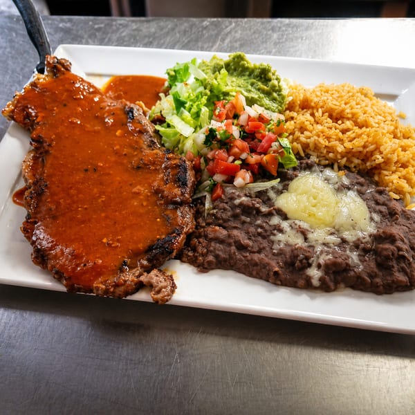 Grilled steak with red sauce, rice, refried beans, guacamole, and pico de gallo on a white plate.