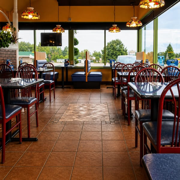 Empty restaurant dining room with red chairs, blue booths, and stained-glass lamps, looking out large windows.