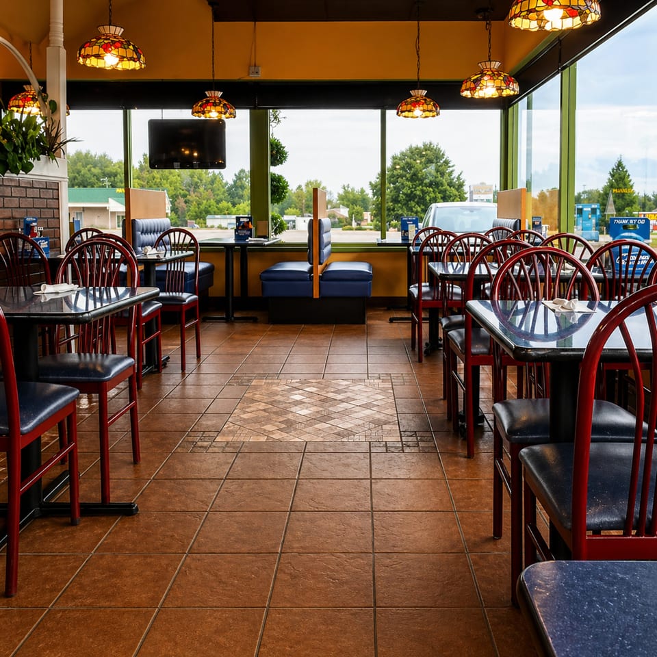 Empty restaurant dining room with red chairs, blue booths, and stained-glass lamps, looking out large windows.