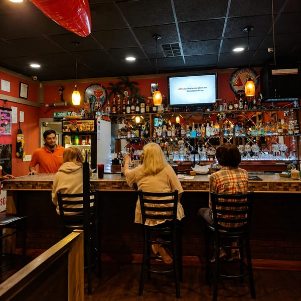 A bartender serves three customers seated at a well-stocked bar with red walls and warm lighting.
