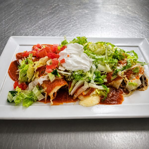 A white rectangular plate holds enchiladas topped with red sauce, shredded lettuce, diced tomatoes, and sour cream.